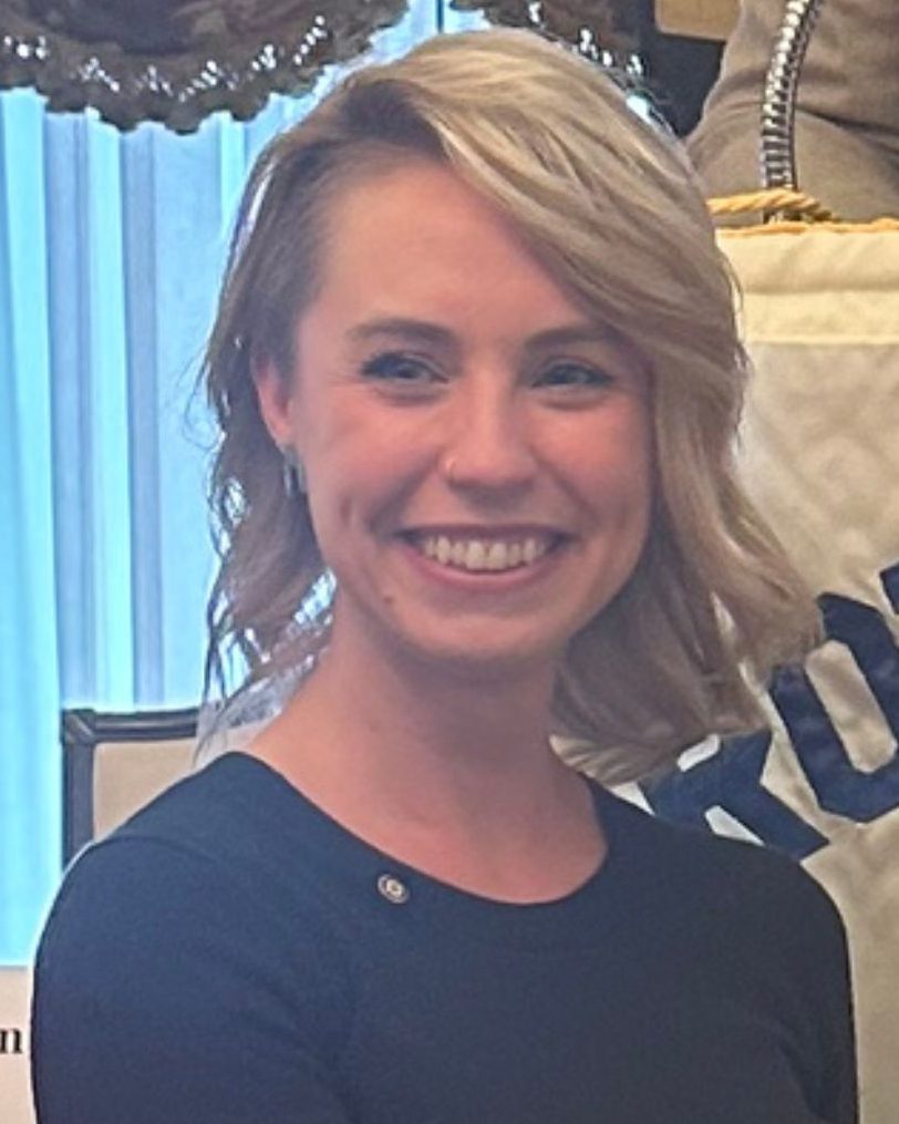 A smiling woman with short blonde hair, wearing a dark top, sitting in a well-lit room with a blue curtain backdrop.